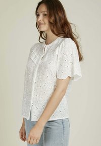 Blouse en coton blanc avec un motif floral brodé, manches courtes à bord scallopé, fermeture à boutons et patte avec plis. Associée à un jean bleu clair.