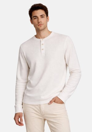 Ung mand iført en hvid langærmet Henley-skjorte med tre knapper og lyse beige bukser, poserende med den ene hånd i lommen.
