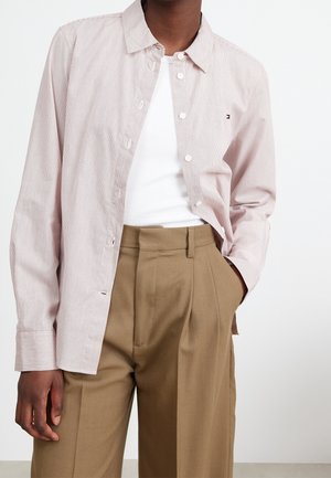 Persona con camisa de botones de rayas rosa claro sobre una camiseta blanca, pantalones marrones holgados de talle alto, con una mano en el bolsillo.