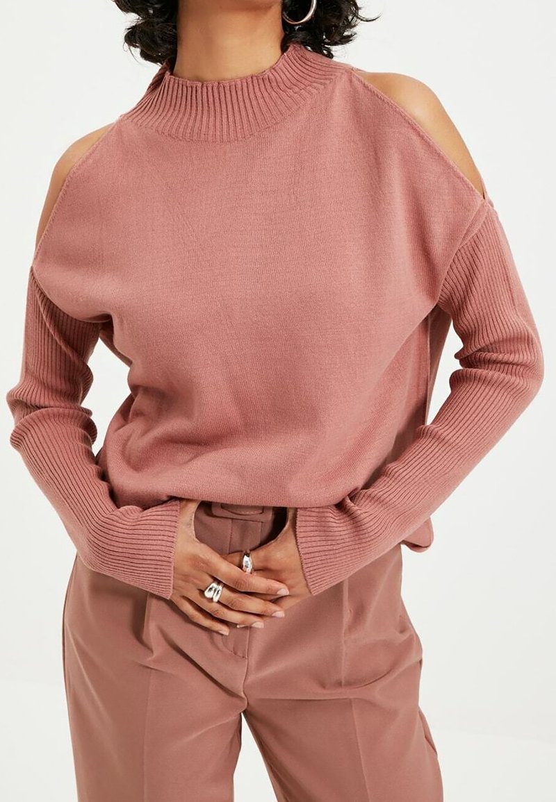 Rosa striktrøje med udskårne skuldre, ribbet høj hals og lange ærmer. Matchet med skræddersyede rosa bukser. Sølvringe på hænderne.