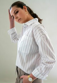 Wit gestreepte shirt met verticale bruine lijnen, button-up kraag, lange mouwen en een voorzak, gecombineerd met beige broeken en een gouden horloge.