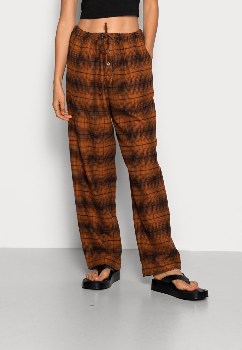 Femme portant un pantalon à carreaux orange et noir avec taille coulissante et des sandales plateformes noires, debout devant un fond uni.