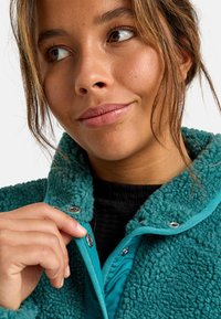 Tegenblauw fleecjasje met een gestructureerde buitenkant, drukknoopsluiting en een gladde binnenvoering, met een hoge kraag en contrasterende stofaccenten.