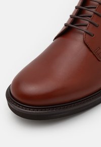 Brune lærstøvle med glat overflade, rund tå og brune snørrebånd. Har en kontrasterende sort sål med syede detaljer.