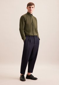 Camicia verde oliva con bottoni e tasca, abbinata a pantaloni blu navy con pieghe e scarpe nere stringate. Tessuto liscio e taglio sartoriale.