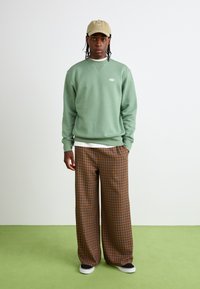 Groene katoenen sweatshirt met ronde hals en wit logo, gecombineerd met bruine, geruite, wijde broek en zwarte sneakers. Bruine pet op het hoofd.