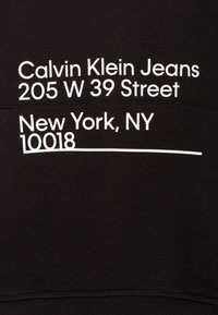 Felpa nera con testo stampato bianco: "Calvin Klein Jeans 205 W 39 Street New York, NY 10018." Morbido misto cotone, polsini a costine.