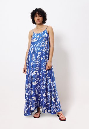 Maxikleid aus blauem Stoff mit weißen Blumenmustern, ausgestattet mit Spaghettiträgern und einem lockeren Schnitt. Kombiniert mit roten, sandalenartigen Riemenschuhen.