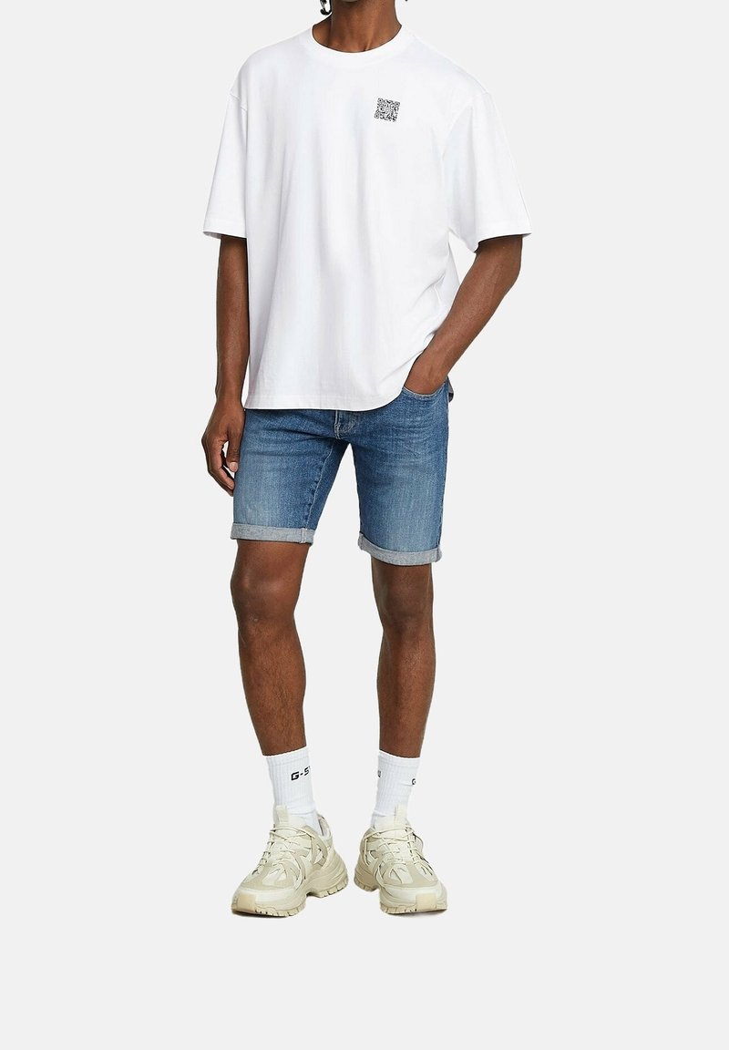 Homme portant un t-shirt blanc oversize avec un petit logo noir, un short en jean bleu retroussé, des chaussettes blanches et des baskets épaisses beiges.