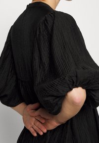 Musta, tekstuuriltaan rikkaita mekko, jossa on puffimaiset hihat ja kerätty vyötärö. Materiaali on ribbikuvioinen ja pehmeä pinta. Kädet ovat ristissä selän takana.
