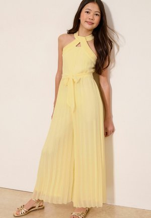 Pige iført en bleg gul halterneck-plisseret jumpsuit med bånd i taljen og metaliske guld-sandaler, stående mod en beige væg.