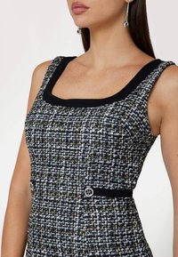 Robe en tweed noir et blanc avec un décolleté carré, tissu texturé et boutons argentés à la taille. Bordure noire lisse sur les épaules.