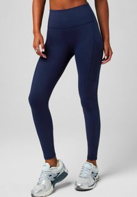 Dunkelblaue, hoch geschnittene Leggings aus dehnbarem Material, mit einer Seitentasche, kombiniert mit silbernen Sportschuhen mit luftiger Oberseite.