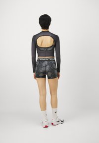 Tumma pitkähihaine paita avoimella selällä, yhdistettynä kuviollisiin mustiin shortseihin. Materiaali näyttää olevan joustava; vyötäröllä on kontrastivärinen logo.