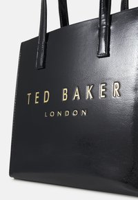 Juodos dirbtinės odos rankinė su dviem rankenomis, su iškiliais auksiniais užrašais "TED BAKER LONDON", išryškintais priekyje.