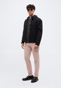 Άνδρας που φοράει μαύρο μπουφάν, σκούρο πουκάμισο, ανοιχτό ροζ παντελόνι, μαύρα δερμάτινα παπούτσια και γυαλιά ηλίου, στέκεται μπροστά σε λευκό φόντο.