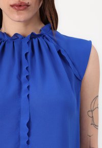 Blauw mouwloos blouse met een rimpelnek en een knopenlijst, gemaakt van soepele stof en met een relaxte pasvorm.
