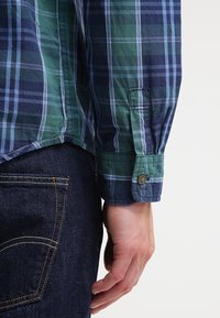 Gros plan sur le bras d’une personne portant une chemise à carreaux bleue et verte à côté d’une poche de jean en denim bleu foncé sur un fond uni.