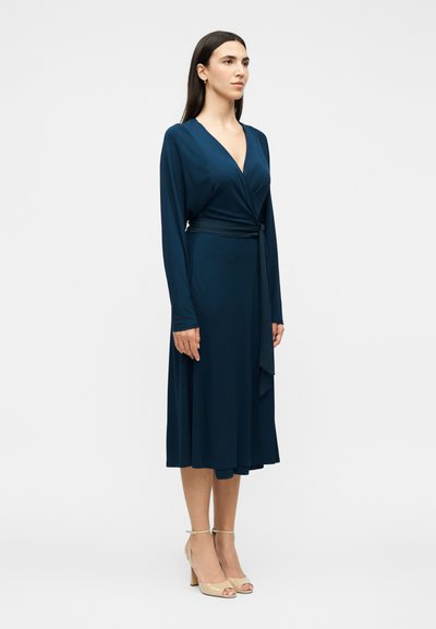 Γυναίκα που φορά ένα μακρυμάνικο, μπλε ρουά φόρεμα τύπου wrap με ζώνη στη μέση, στέκεται με μπεζ σανδάλια με ψηλό τακούνι σε ουδέτερο φόντο.