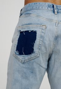 Lyseblå denimjeans har et slitt design med en stor lapp av mørkeblått stoff på baklommen, og viser frynsete kanter.