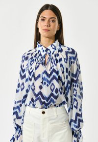 Langærmet blouse i let, mønstret stof med blå og hvide zigzag- og prikkemønstre; har en bindebånd i halsen og voluminøse ærmer.