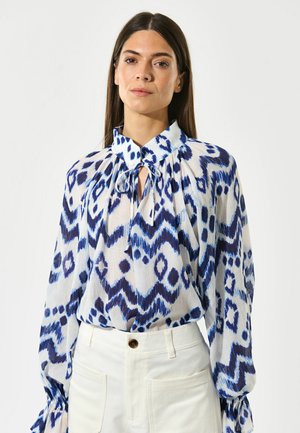 Långärmad blus i lätt, mönstrad tyg med blå och vita zigzag- och prickmönster; har en knytkrage och voluminösa ärmar.
