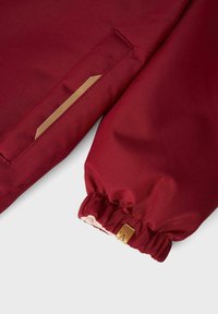 Süva maroon värvi jope varrukas, millel on elastne mansett ja väike nahklabel, millel on osaliselt nähtav õmmeldud tasku.
