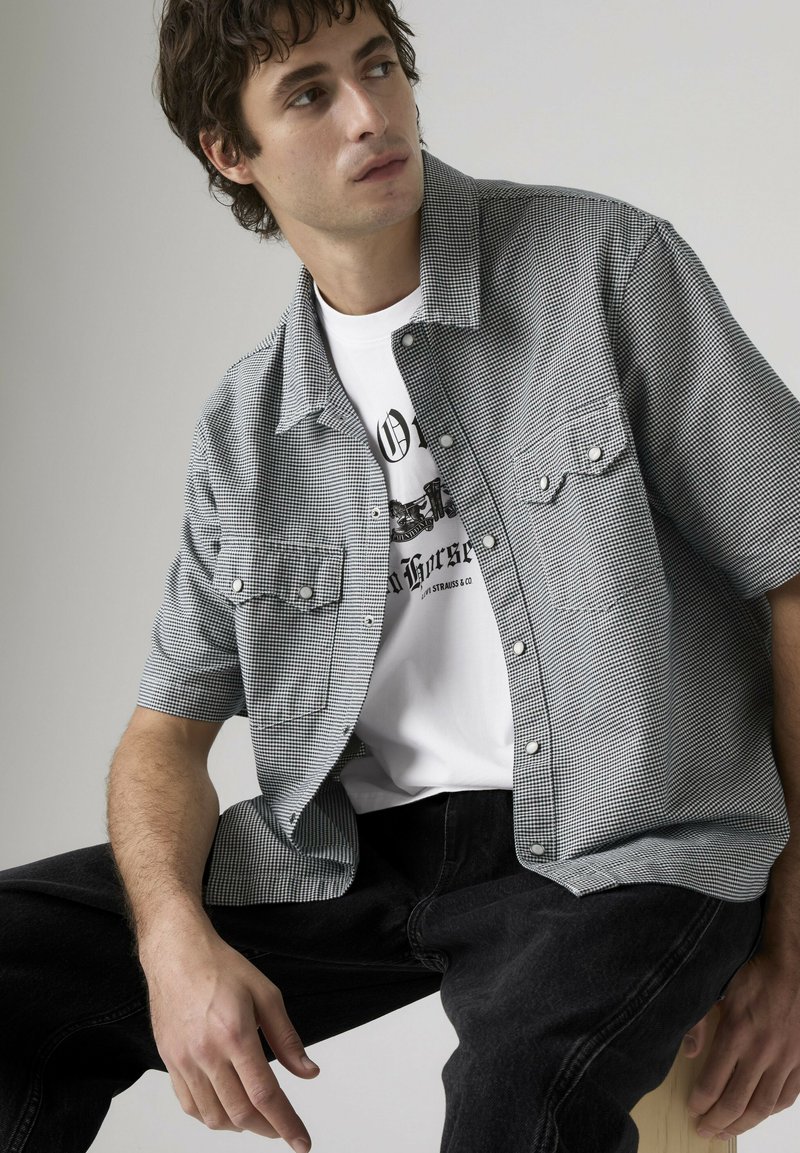 Giovane uomo con capelli ricci che indossa una camicia a maniche corte a quadri sopra una t-shirt bianca con stampa, seduto e che guarda verso sinistra.