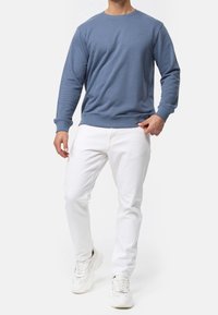 Homme portant un sweat à manches longues bleu, un jean blanc et des baskets blanches, se tenant devant un fond clair uni.