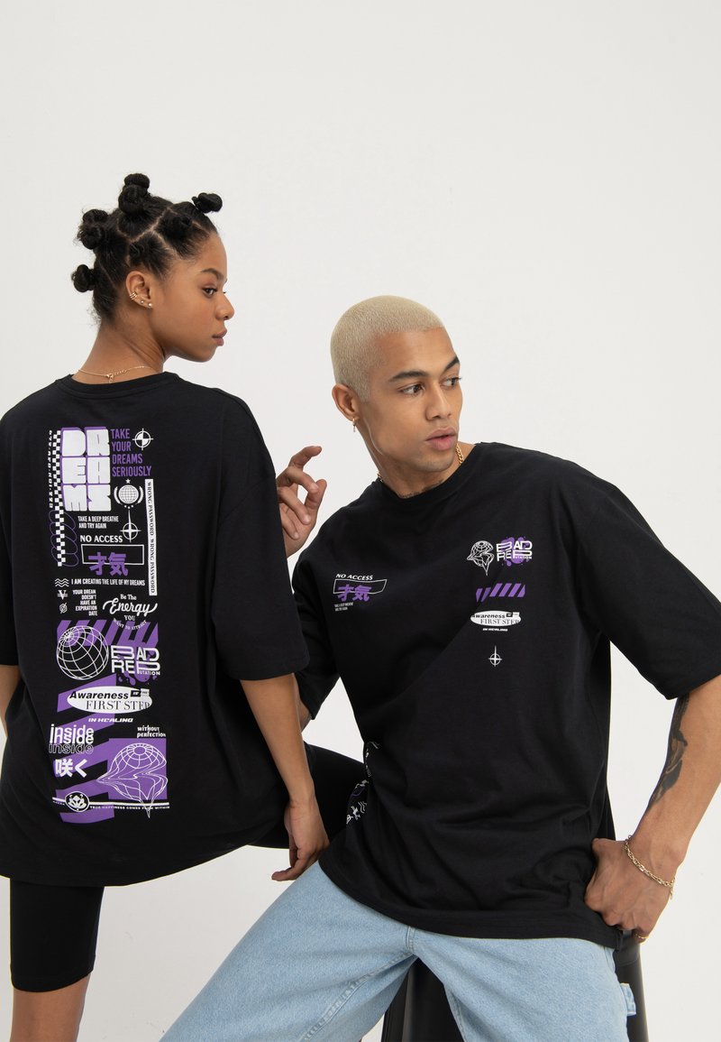 To modeller iført svarte grafiske t-skjorter med lilla og hvit tekst og design, poserer foran en enkel hvit bakgrunn.