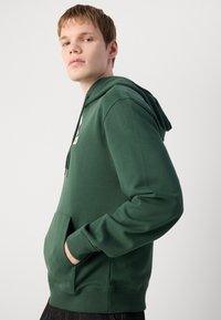 Felpa verde con tasca a kangaroo, cappuccio con coulisse e polsini a costine. Presenta una piccola patch con logo sul petto e una texture morbida.