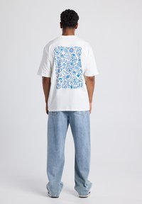 White cotton t-shirt featuring a blue graphic design on the back, depicting various abstract shapes and elements. Paired with light blue jeans.