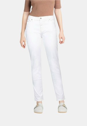 Donna che indossa jeans bianchi a gamba dritta con bottone e cerniera, abbinati a scarpe slip-on bianche e top marrone a coste.