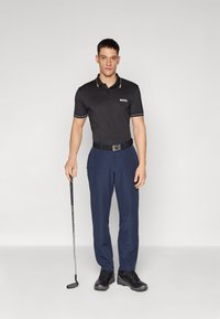 Polo negro con detalles dorados y logo, pantalones azul marino, cinturón y zapatos negros; de pie con un palo de golf sobre un fondo simple.