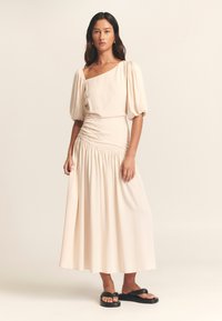Beige jurk van gemiddelde lengte met pofmouwen en een één-schouder ontwerp. Heeft geplooide details bij de taille. Gecombineerd met zwarte platte sandalen.