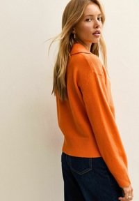 Maglione lavorato a maglia arancione con colletto, caratterizzato da una vestibilità rilassata e orlo a coste. Abbinato a jeans di denim blu scuro. Tessuto morbido.