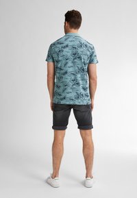 Polo tričko s tyrkysovým vzorem a tmavým květinovým designem, doplněné o zahnouté tmavé šortky a bílé tenisky. Pozadí je jednobarevné.