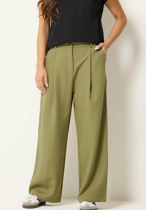 Vrouw die olijfgroene wijde broek, zwarte top en witte sneakers draagt, staand met één hand in haar zak tegen een lichte achtergrond.
