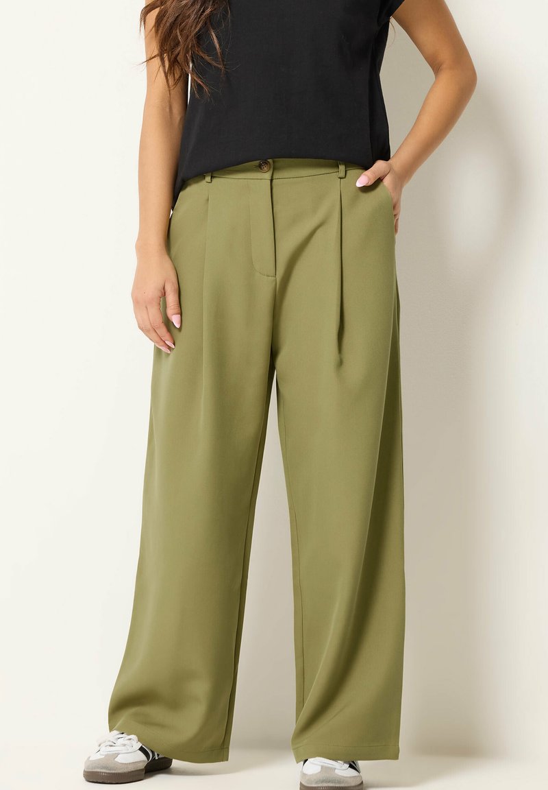 Femme portant un pantalon large vert olive, un haut noir et des baskets blanches, debout avec une main dans la poche sur un fond clair.