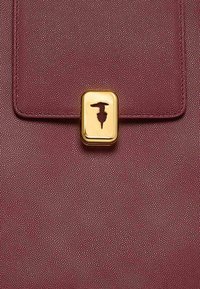 Zbliżenie na bordową, teksturowaną skórzaną torbę z przednią klapką i złotym, prostokątnym zapięciem z małą wytłoczoną postacią.