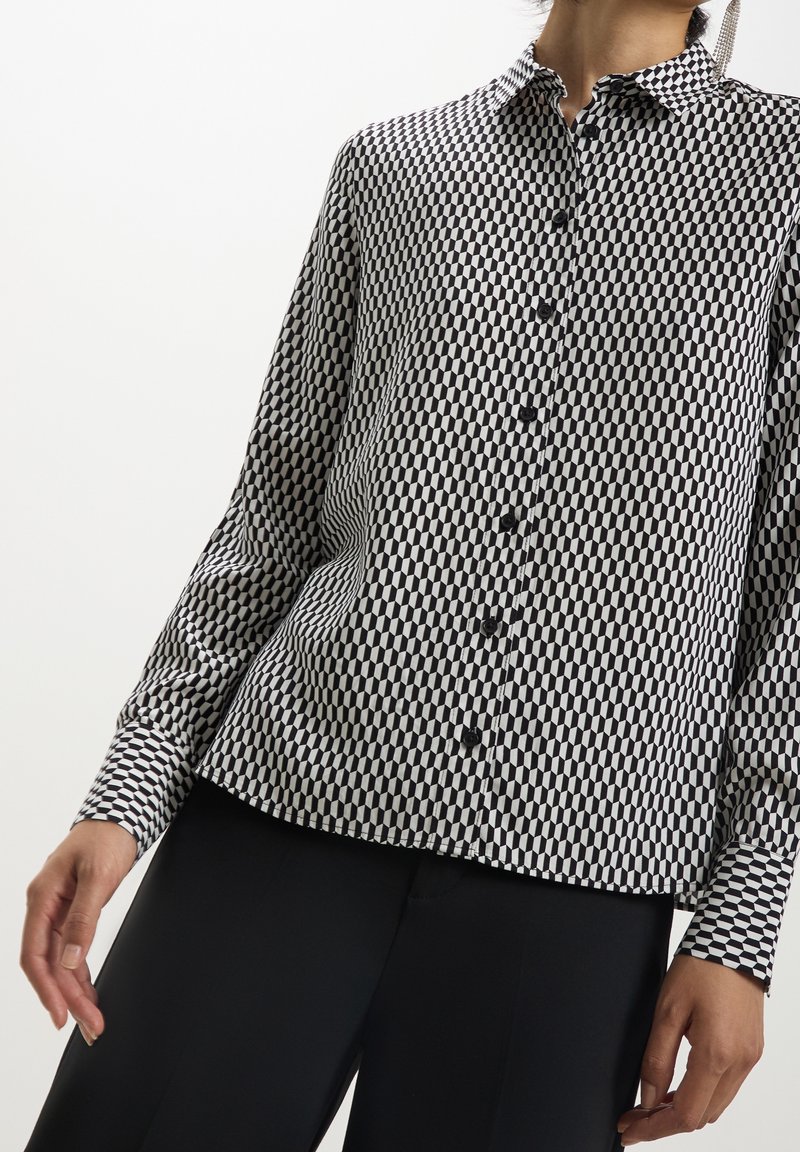 Chemise à manches longues boutonnée avec motif géométrique noir et blanc, portée avec un pantalon noir, montrant le torse et partiellement les bras.