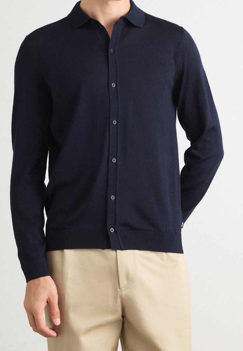 Homme portant un cardigan bleu marine boutonné et un pantalon beige, debout devant un fond clair uni avec une main derrière le dos.