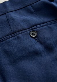 Nahaufnahme eines marineblauen Stoffes mit strukturierter Webart, der einen schwarzen Vierloch-Knopf und eine detailreiche, genähte Naht an einem Kleidungsstück zeigt.