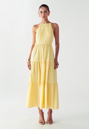 Yellow halter-neck maxi dress with tiered ruffles, smooth texture, and fitted waist. Worn with neutral heeled sandals.