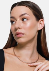 Metallkette mit drei dünnen, geschwungenen Strängen, die eine glatte, glänzende Goldoberfläche aufweist, getragen von einem Modell mit glattem, langem Haar.