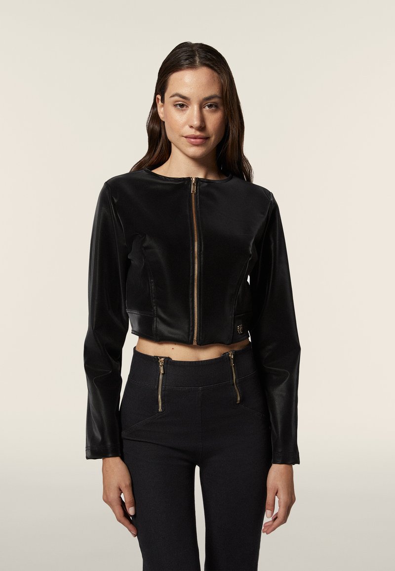 Giacca nera corta con una texture liscia, scollo rotondo, zip frontale e maniche lunghe. Abbinata a pantaloni neri a vita alta con zip.