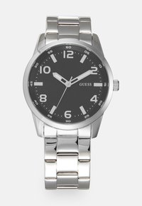 Silberner Edelstahl-Armbanduhr mit einem runden schwarzen Zifferblatt, weißen Stundenmarkierungen und einem Metallgliederarmband; enthält ein Logo in der Nähe der Mitte.