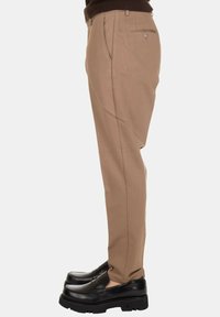 Persona con pantaloni beige su misura dotati di tasche laterali e posteriori, abbinati a mocassini neri spessi, in piedi contro uno sfondo chiaro e semplice.