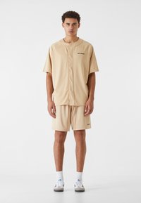 T-shirt de sport en maille beige à manches courtes, présentant un col rond et un bouton à l'avant. Accompagné de shorts assortis, tous deux avec un branding discret.
