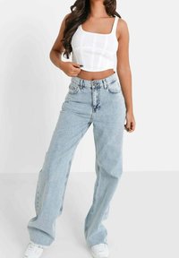 Donna che indossa un crop top bianco aderente e jeans a vita alta leggeri e larghi di colore blu chiaro, con scarpe da ginnastica bianche, in piedi di fronte a uno sfondo semplice.